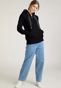 Schwarzer Hoodie mit Fronttaschen und Kordelzug, kombiniert mit hellblauen, lässig sitzenden Jeans und weißen Sneakern auf einem neutralen Hintergrund.