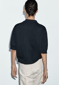 Maglione polo lavorato a maglia nera con maniche corte, colletto a coste e orlo. Indossato con pantaloni chiari. La vista posteriore mostra dettagli di design minimali.