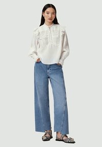 Blusa blanca con mangas acampanadas, pliegues detallados y botones, combinada con unos vaqueros de pernera ancha azul y sandalias negras con estampado.