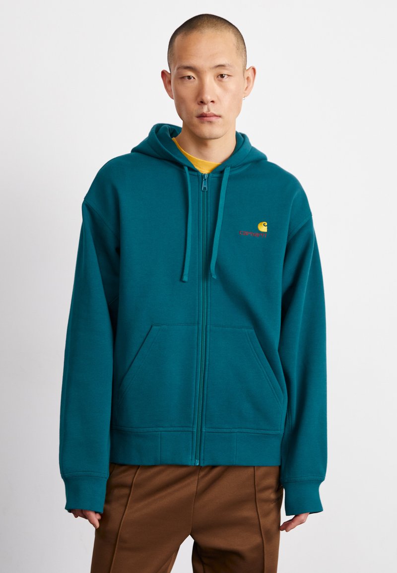 Mėlynos spalvos zip-up gobtuvė, pagaminta iš švelnaus audinio, turinti priekinę kišenę, raištelius gobtuve ir siuvinėtą logotipą geltonos ir raudonos spalvų krūtinėje.