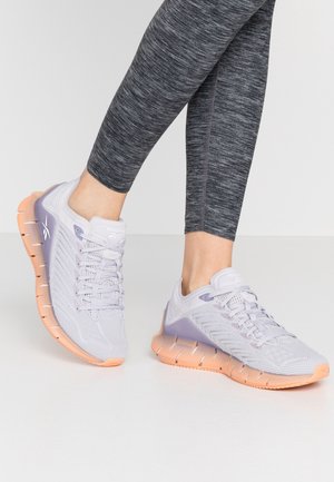 Person iført grå leggings og hvide og ferskenfarvede Reebok træningssko på en lys baggrund.