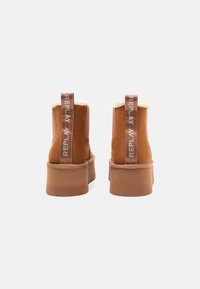 Pár hnědých semišových platformových kotníkových bot s krémovou kožešinou uvnitř a značkovými poutky na zadní straně, zobrazeno zezadu na bílém pozadí.