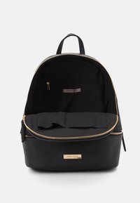 Sac à dos noir avec une forme arrondie, en tissu texturé, fermeture éclair dorée et poches intérieures. Étiquette de logo à l'intérieur. Design minimaliste.