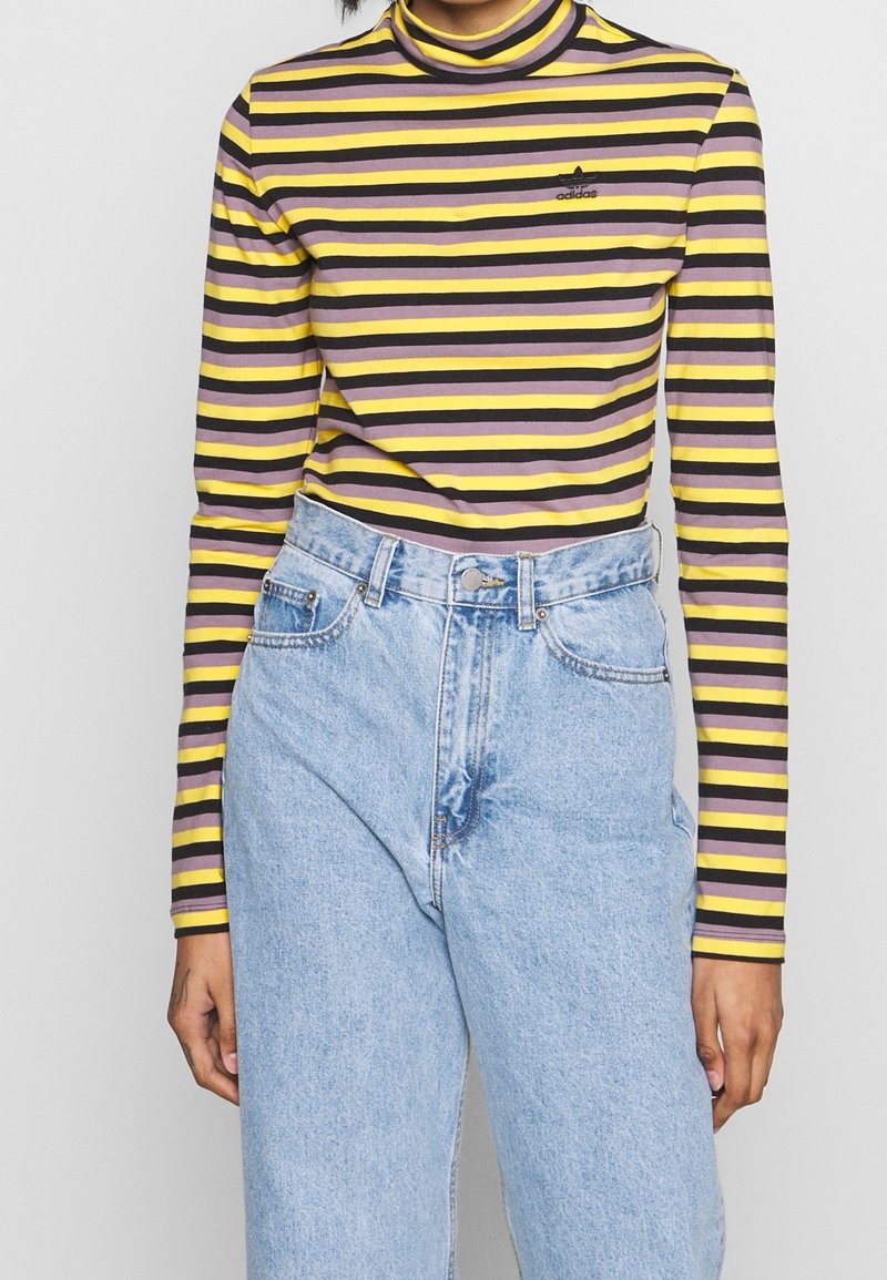 Chemise à rayures à manches longues jaune, violet et noir avec un col haut, associée à un jean clair taille haute. Logo Adidas visible.