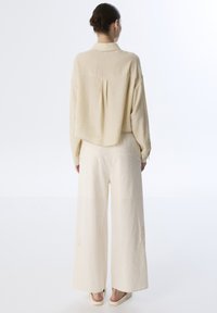 Camisa oversized de color beige con textura, cuello y pliegue en la espalda. Combinada con pantalones anchos de color claro con bordados florales sutiles.