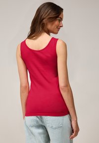 Top sin mangas ajustado de color rojo con escote redondeado y textura suave, combinado con jeans de mezclilla azul claros. La parte trasera presenta un diseño simple y limpio.