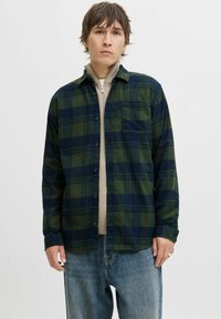 Junger Mann, der ein grün und marineblau kariertes Hemd über einem beigen Reißverschlusspullover und lockeren blauen Jeans trägt, vor einem schlichten Hintergrund steht.