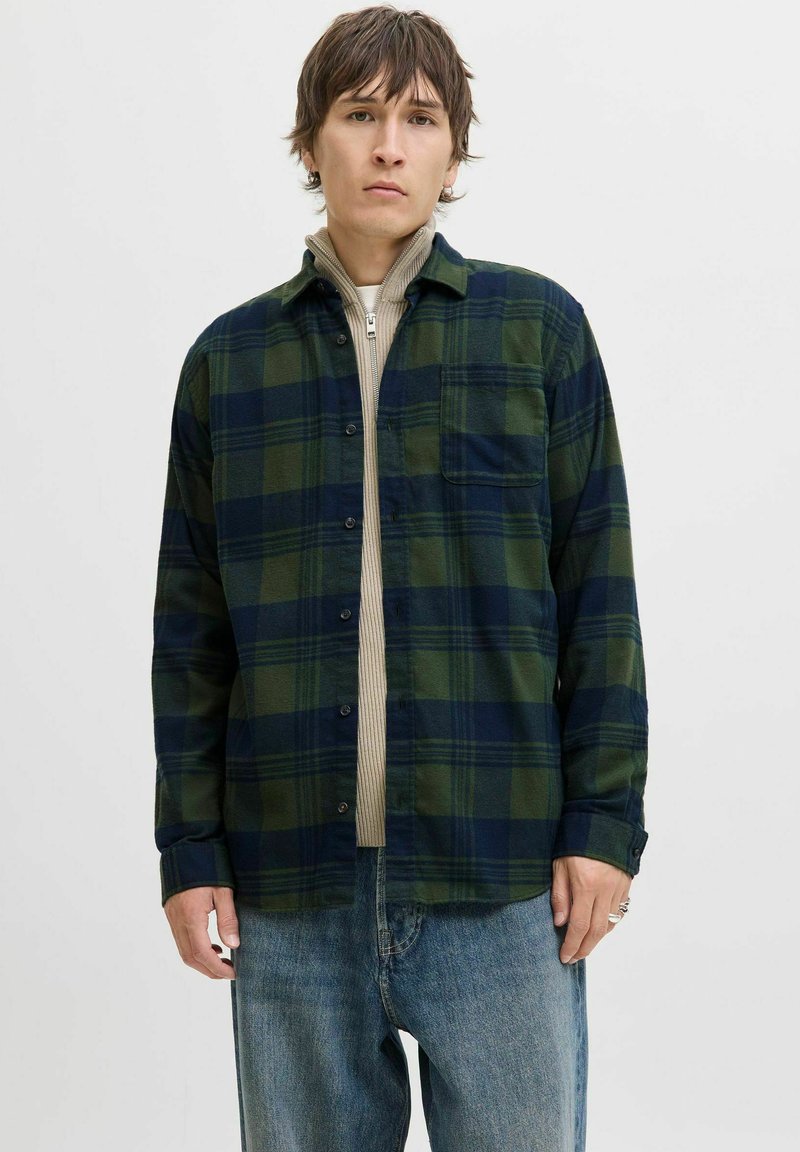 Junger Mann, der ein grün und marineblau kariertes Hemd über einem beigen Reißverschlusspullover und lockeren blauen Jeans trägt, vor einem schlichten Hintergrund steht.