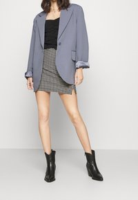 Šedý oversized blazer s saténovými manžetami, černý nabíraný top a kostkovaná minisukně se skladem, doplněné černými kotníkovými botami.