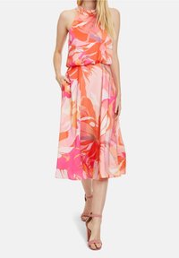 Kvinna som bär en ärmlös, höghalstrad midi-klänning i rosa och orange med blommönster och fickor, samt rosa klacksandaler, går framåt.