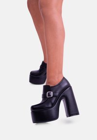 Scarpe platform in faux leather nere con punta arrotondata, alto tacco a blocco e una fascia con fibbia sul collo del piede. Texture liscia e design elegante.