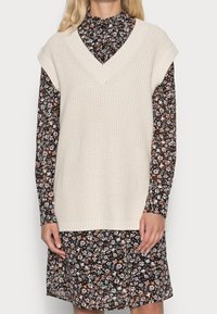 Pletená krémová vesta s výstřihem do V a širokými průramky, vrstvená přes černé květinové šaty s dlouhými rukávy a vysokým límcem.