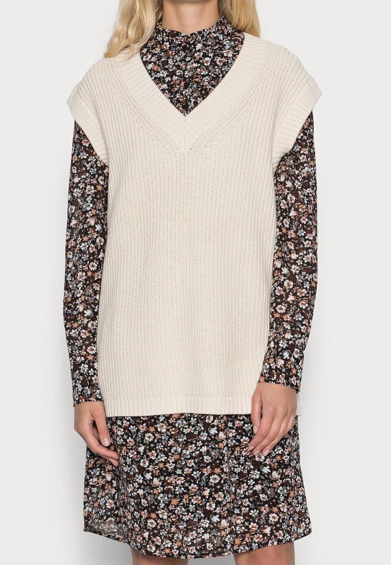 Pletená krémová vesta s výstřihem do V a širokými průramky, vrstvená přes černé květinové šaty s dlouhými rukávy a vysokým límcem.
