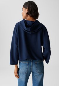 Donna con capelli corti e scuri, vestita con un top con cappuccio texturato di colore blu navy e jeans di denim blu, in piedi con la schiena rivolta verso la macchina fotografica.