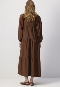 Bruine, gelaagde maxi-jurk met lange puffmouwen en een getailleerde bovenkant. Gemaakt van zachte stof, met een vloeiende silhoutte en geplooide accenten.