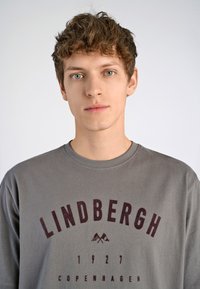 T-shirt gris avec un col rond et "LINDBERGH COPENHAGEN 1927" imprimé en bordeaux. Tissu doux avec une coupe décontractée.