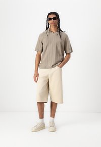 Camisa polo de manga corta beige con relieve y escote en V, combinada con pantalones cortos beige claros hasta la rodilla y zapatillas de color crema.
