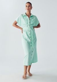 Abito button-down verde menta con maniche corte, due tasche sul petto, vita con cintura e orlo di lunghezza media. Tessuto liscio con una leggera texture.