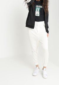 Svart zip-up jacka med vita ränder, svart t-shirt med grön branding, vita joggerbyxor och vita sneakers, allt i en avslappnad sportig stil.