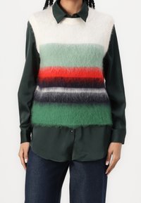 Sweter bez rękawów w zielone, czerwone i granatowe paski, noszony na ciemnozielonej koszuli z guzikami. Wykonany z miękkiego, puszystego materiału o luźnym kroju.