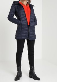 Marinblå vadderad jacka med huva och dragkedja, med sidofickor. Matchad med en orange stickad tröja, svarta tajta jeans och svarta stövlar.