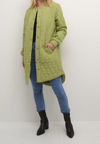 Person klædt i en lang lysegrøn quiltet frakke med trykknapper, blå stramme jeans og sorte hælede ankelstøvler på en hvid baggrund.