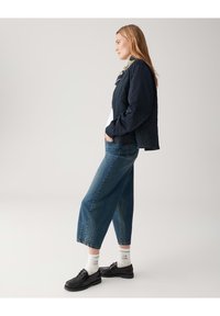 Navy quilted jacket, relaxed-fit light denim crop pants, black shoes, and white socks with green text. Simple, functional design and colors.