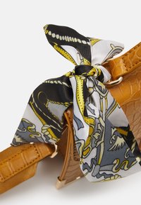 Foulard en soie à motif noir, jaune et gris noué à la bandoulière d'un sac à main couleur sable avec texture crocodile et ferrures dorées.