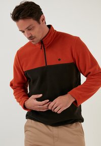 Fleece pullover featuring an orange top section and black lower section, with a quarter zip and small logo on the chest. Soft texture.