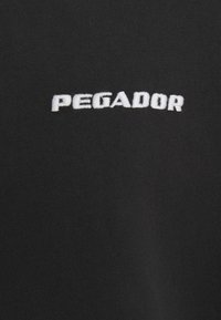 Svart t-skjorte i stoff med hvit brodert tekst "PEGADOR" sentrert på brystet. Glatt tekstur med standard rund hals.
