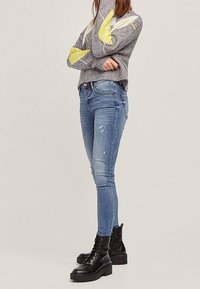 Grijze trui met gele en witte patronen, gecombineerd met blauwe skinny jeans met verweerde details en zwarte veterlaarsjes tot aan de enkel.