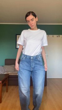 Jong persoon met kort haar, gekleed in een wit T-shirt en losse blauwe spijkerbroek, staand in een woonkamer met groene muren.