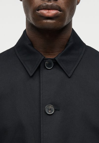 Κοντινό πλάνο ενός σκούρου δέρματος προσώπου που φοράει μαύρο παλτό με γιακά, με δύο ορατά μαύρα κουμπιά κλεισμένα.