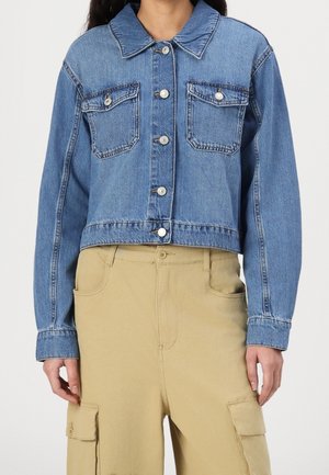 Kobieta ubrana w krótką niebieską kurtkę dżinsową z zapinanymi na guziki kieszeniami na piersi oraz beżowe spodnie typu cargo z bocznymi kieszeniami.