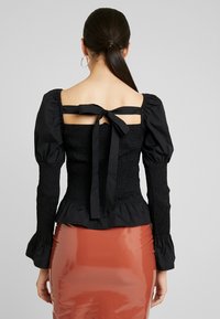Svart topp med puffärmar i smockat tyg, med en fyrkantig rygg och knytning. Matchas med en glansig kjol i rostfärg.