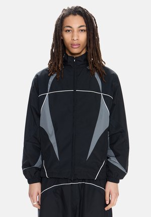 Nuori henkilö, jolla on pitkät rastat, pukeutuneena musta-harmaaseen vetoketjulliseen urheilutakkiin, jossa on joustavat hihansuut, seisomassa yksinkertaisen taustan edessä.