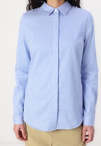 Chemise bleu clair à boutons avec de fines rayures verticales blanches, design à col, manches longues et ourlet arrondi. Le tissu semble lisse.