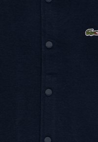 Cardigan blu navy con bottoni in plastica, realizzato in un tessuto a maglia texturizzata. Presenta un logo di coccodrillo ricamato sul lato sinistro.