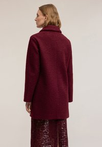 Vínový kabát s texturou, klopovým límcem, dlouhými rukávy a střední délkou, doplněný krajkovou sukní pod ním.