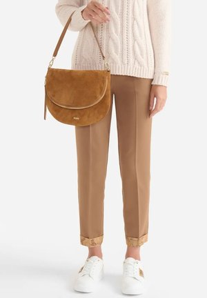 Borsa a tracolla in camoscio color cuoio con chiusura a patta, dettagli in oro e tasca con cerniera. Indossata con pantaloni beige su misura e sneakers bianche.