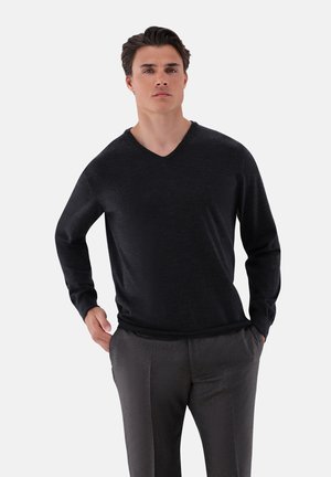 Schwarzer V-Ausschnittpullover mit langen Ärmeln, aus einem glatten, weichen Stoff. Das Model trägt graue, maßgeschneiderte Hosen mit einer lockeren Passform.