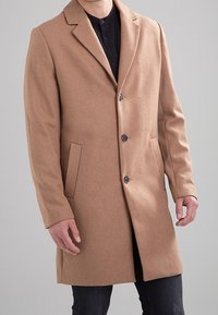 Homme portant un manteau en laine camel boutonné par-dessus une chemise noire et un jean foncé, se tenant devant un fond gris clair uni.