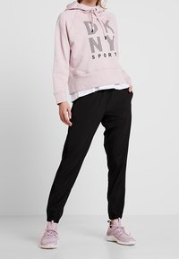 Sweat à capuche rose clair avec "DKNY SPORT" imprimé en noir ; associé à un pantalon de jogging noir et des chaussures de sport roses claires.