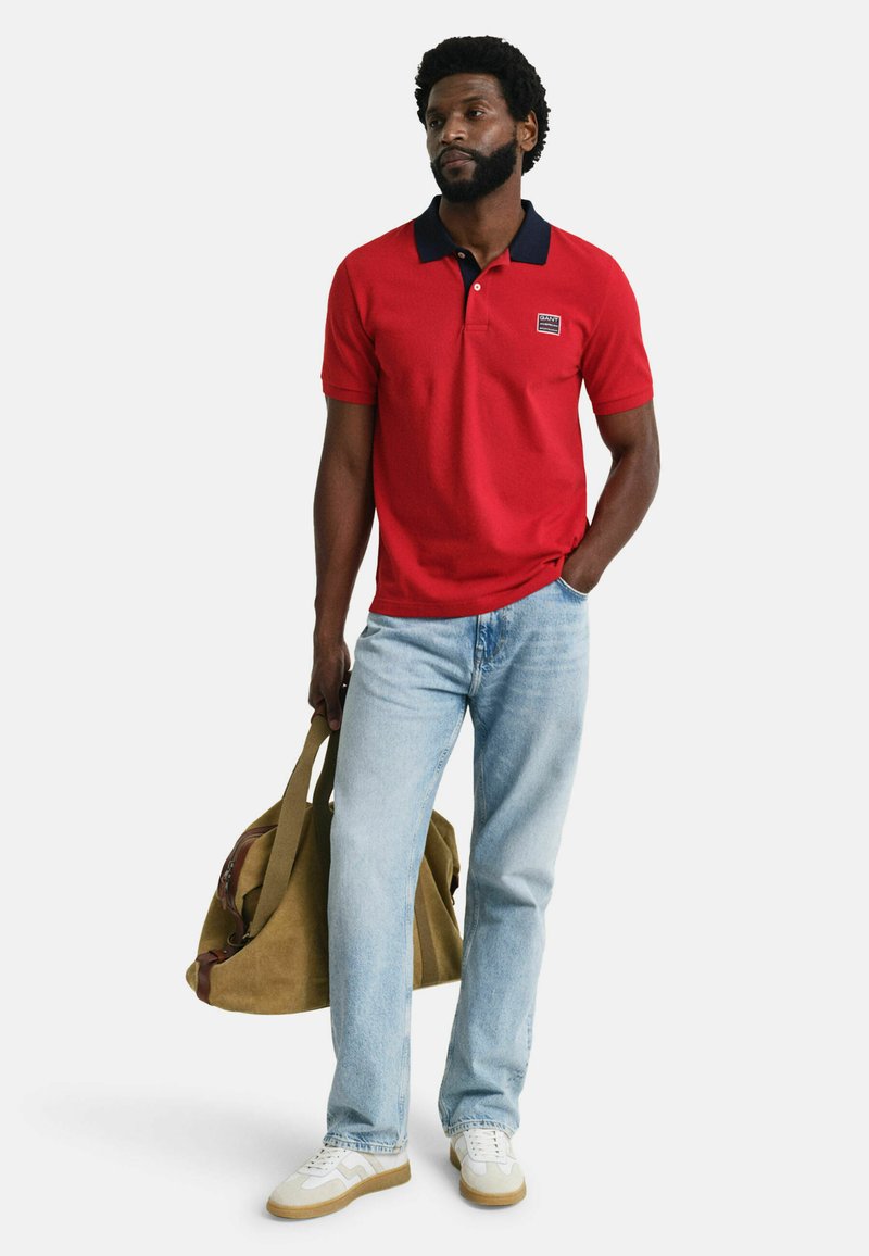Rotes Poloshirt mit schwarzem Kragen, hellblaue Jeans und weiße Sneakers. Hält eine braune Canvas-Reisetasche mit Lederakzenten.