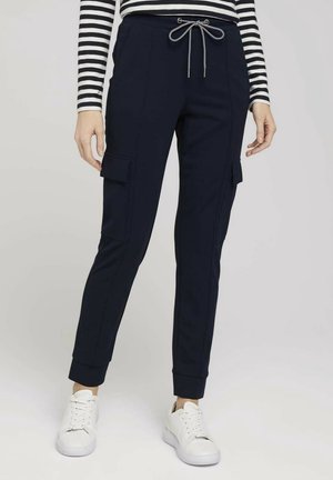 Person iført navy cargobukser med snøre i taljen, stribet langærmet top og hvide sneakers, stående på ensfarvet baggrund.