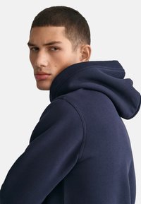 Felpa blu navy in tessuto testurizzato, con un grande cappuccio regolabile e polsini a costine. Il modello guarda oltre la spalla.