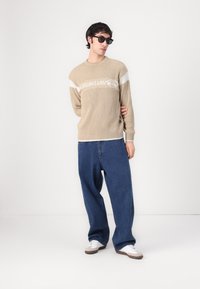 Beige gerippter Pullover mit weißem Logo-Detail, kombiniert mit lockeren blauen Jeans. Das Model trägt schwarze Sonnenbrillen und weiße Sneakers.