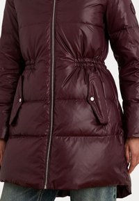 Burgundijas pūkains mētelis ar sasprindzinātu vidukli, ar rāvējslēdzēju un divām sānu kabatām ar pogām. Gluda, spīdīga auduma ar quilted rakstu.