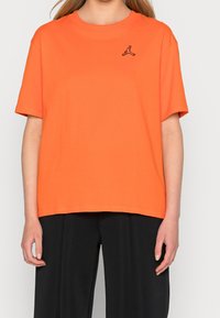 Pomarańczowy bawełniany t-shirt z krótkimi rękawami i okrągłym dekoltem. Na lewej piersi znajduje się małe czarne logo. Prosty dół koszulki.