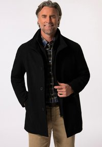 Abrigo de mezcla de lana negro con cuello alto, presenta dos botones frontales y bolsillos laterales, superpuesto a una camisa de cuadros, llevado por un hombre sonriente.